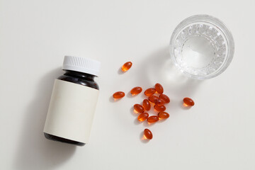 On a white background, there is a glass of water, an unlabeled medicine bottle, and a few orange soft capsules. A scene mimicking the advertising of health-supporting functional food.