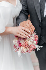 Obraz na płótnie Canvas The hands of the bride and groom with rings on the background of a wedding bouquet of white-pink flowers and dried flowers