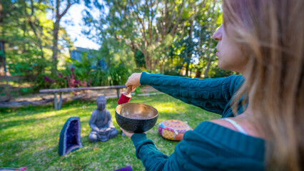 Young woman with a singing Tibetan bowl in a garden. Buddha statue and amethyst stones in the...