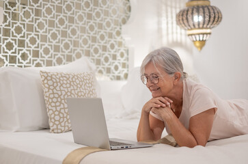Smiling senior woman at bedroom at home or hotel room looking at laptop enjoying tech and social. Elderly female online with computer