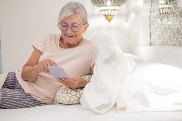 Smiling senior woman lying in bedroom at home or hotel room looking at mobile phone enjoying tech and social. Elderly female online typing on smart phone
