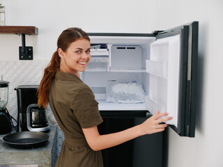 Fototapeta na wymiar Woman smiling with teeth looking into camera in kitchen at home opened freezer empty with ice inside, home refrigerator, defrosted, view from back.