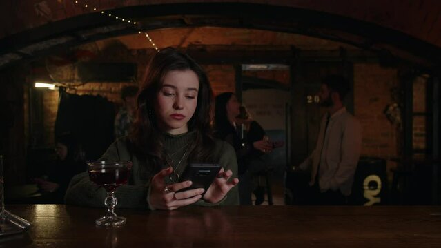 Alone beautiful girl browsing social media in bar counter. Young pretty woman using smartphone at the terrace cafeteria. Summer evening with favorite cocktail. Lonely girlfriend texting friends