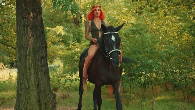 Sexy fantasy woman queen forest nymph sits on horseback, rides black horse pet animal. Fairy tale girl. elf dress orange red long hair wreath diadem flowers, summer nature forest green trees leaves