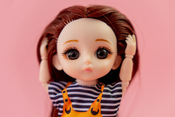 child toy doll with dark hair in an orange dress standing on a pink background. Plastic children's toy. Doll games for imagination. the frightened doll holding his head in fear.