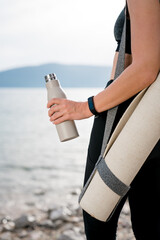 Reusable water bottle and yoga mat in woman hand. Sport, wellness on sea beach. Girl resting after...