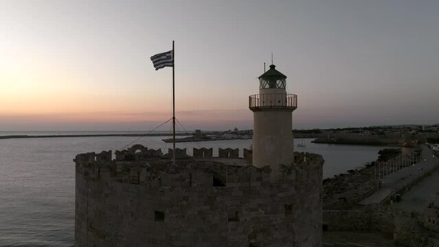Sunrise over Medieval fortification lighthouse and Greek flag in the island of Rhodes, Greece