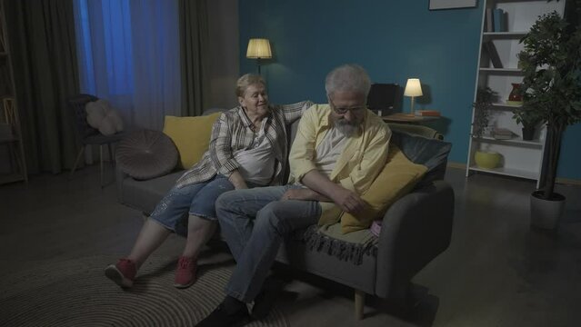 In the picture the elderly couple sit on the sofa in the apartment against the blue wall. The man gives a gift to the woman. She is surprised, excited and delighted. Romance. HDR BT2020 HLG Material.