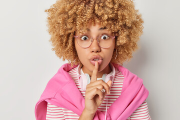 Photo of beautiful curly haired woman makes silence gesture keeps index finger over lips asks to be...