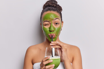 Horizontal shot of dark haired Latin woman drinks fresh smoothie applies nourishing green facial mask made on natural products wrapped in bath towel isolated over white bacground. Detox concept
