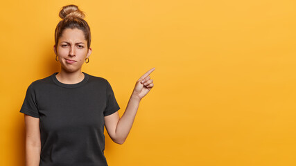 Waist up shot of discontent sulking woman frowns face indicates at blank copy space with frustrated expression dressed in casual black t shirt isolated on yellow background. Advertisement concept - obrazy, fototapety, plakaty