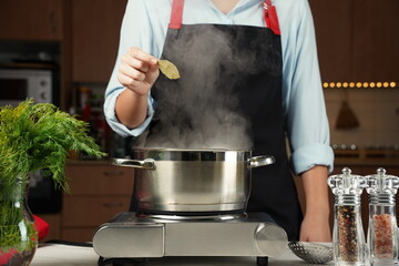 A girl adds bay leaf in steel cooking pan on electric hob with boiling water