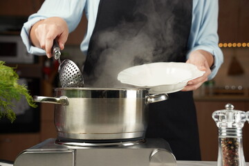 Woman with a ladle and a plate in hands. Steel cooking pan on electric hob. The process of cooking dumplings