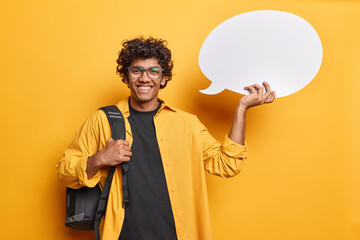 Waist up shot of cheerful Hindu student poses with communication bubble carries rucksack suggests to write your ideas here has glad expression wears shirt isolated over yellow studio background - Powered by Adobe
