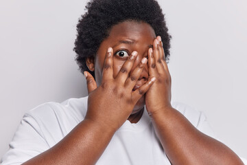 Shocked scared overweight young African woman with curly hair covers face with hands peeks through fingers being scared of something dressed in casual t shirt isolated over white background.