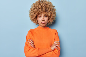 Horizontal shot of displeased young woman with fair curly hair keeps arms folded has offended upset expression wears casual orange jumper isolated over blue background. Negative emotions concept - obrazy, fototapety, plakaty