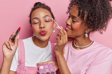 Photo of curly haired woman applies eyeshadow to friend do makeup together put on lipstick prepare...