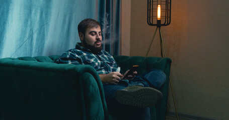 man vaping on a green armchair , man checking social media via phone. man exhaling vapor from an electronic cigarette. 