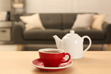 Red cup of fresh tea and teapot on wooden table in room