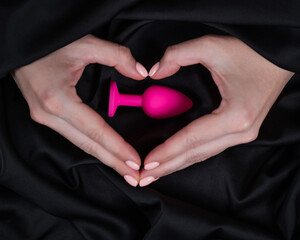 Woman making a heart shape with her palms around a pink butt plug on a black silk sheet. 