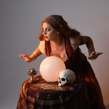 close up portrait of beautiful red haired woman wearing a medieval fantasy fortune teller costume, looking into crystal ball reading the future at seance table. isolated on studio background 