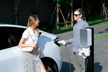 Young couple travel with EV electric car charging in green sustainable city outdoor garden in...