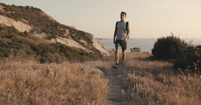 Hiker man in a T-shirt and shorts with backpack walk on the coast with warm sun light.
