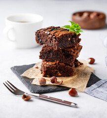 A stack of chocolate brownies with mint, hazelnuts and salted caramel on baking paper on a light background with a cup of coffee.
