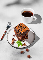 A stack of chocolate brownies with mint, hazelnuts and salted caramel on a white plate on a light background with cup of coffee and morning shadows.