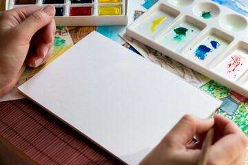 Woman hands, sketchbook - white sheet (workspace), different colors of watercolors in the palette