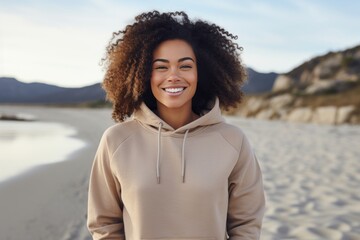 an advertisement lifestyle photo of a gorgeous young sporty african woman in the beach wearing a beige colored empty hoodie. Clothes mock-up for brand placement. Product design template