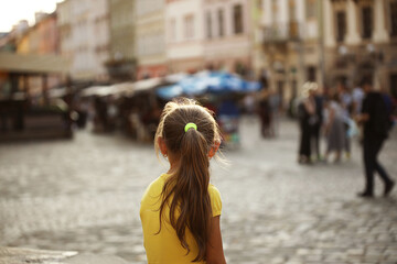 A little girl blonde looks into the distance standing on the street of a medieval European city....
