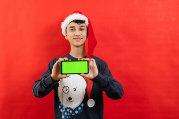 Portrait of a young guy in christmas outfit and santa hat on red background, holding smartphone with green screen