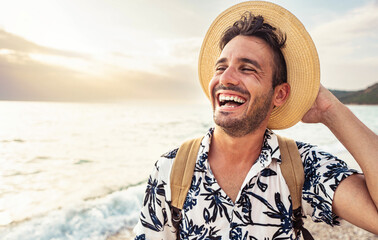 Portrait of cheerful caucasian young man with hat and backpack enjoying sunset at the beach - Laughing guy having fun outside - Well being, healthy life style and traveling concept - obrazy, fototapety, plakaty