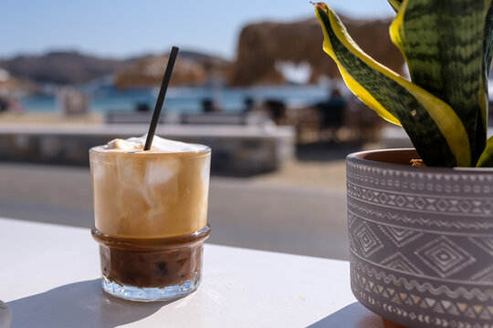 View of an iced Greek  coffee, also known as freddo cappuccino with a straw and the beach of Gialos in Ios Greece