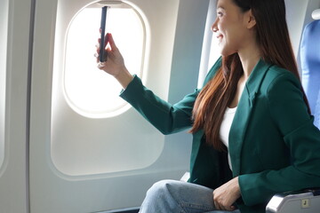 Asian woman sitting on airplane playing with cell phone and looking out window on vacation travel...
