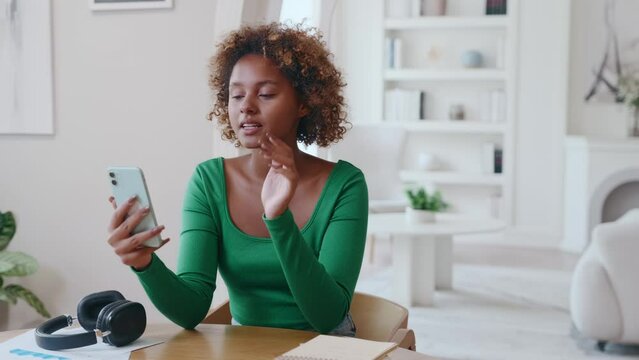 Young beautiful sociable African American woman with phone waves hand and says hello starting video call in messenger or recording short movie for social network sits at table in apartment.