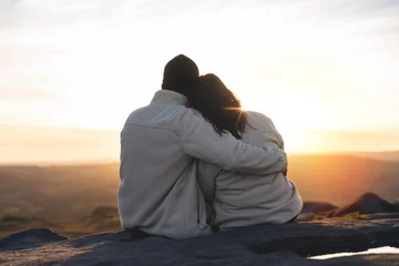 Zelfklevend Fotobehang Back view of the happy couple in love sitting on top of a mountain enjoying a sunset landscape view © Iryna