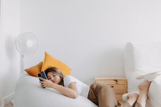 Unhappy Asian Thai woman lying on bean bag sofa, spending time alone at apartment room lonely, holding and using phone waiting for boyfriend texting. get bored and lazy.