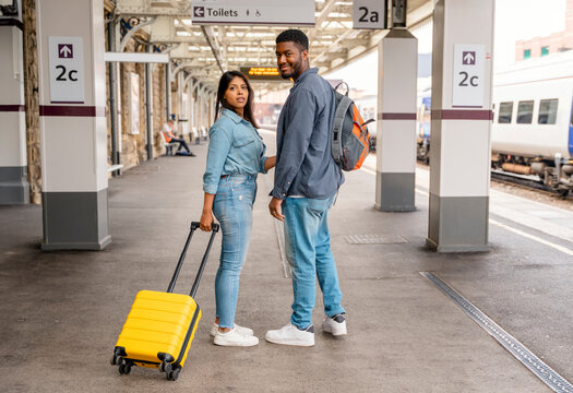 Happy loving couple with luggage and backpack walking along railway platform as they are ready to travel on holiday. Man and woman holding  hands,  looking back and smiling. Travel by train concept