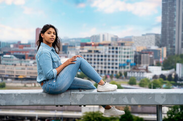 Fototapeta premium Portrait of a beautiful Indian woman sitting on the barrier in front of a modern city background dreaming about the future