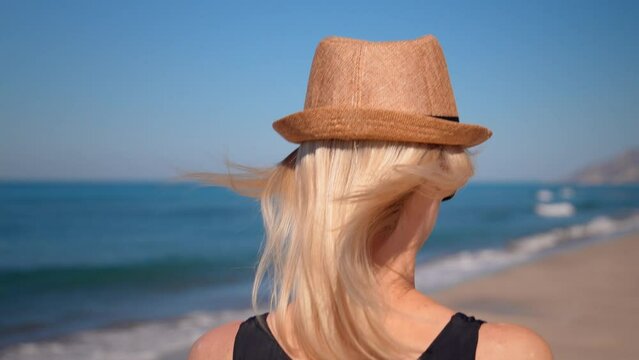 Close-up view of young woman in sea. Girl walks on beach with sea and waves along shore overlooking mountains. Female view on sunny hot day and walking in hat on beach.