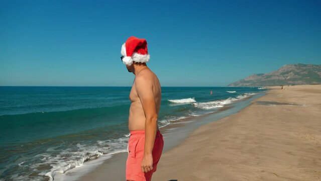 Close-up of man, guy in santa claus hat at christmas or new year on seashore on beach on background of mountains in sunglasses. walks along shore with view of mountains. Journey to Holiday.