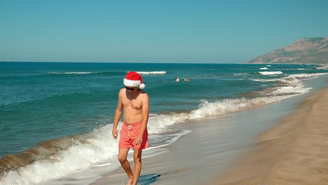 Close-up of man, guy in santa claus hat at christmas or new year on seashore with waves on beach on background of mountains in sunglasses. walks along shore with view of mountains. Journey to Holiday.