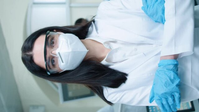 Vertical orientation of beautiful female pharmacist or biologist with respirator and goggles looking seriously at camera. Smart woman wearing gloves and crossing hands while standing in laboratory.