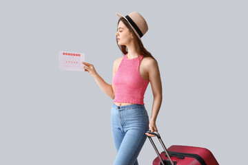 Young woman with menstrual calendar and suitcase on light background