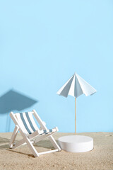 Creative composition with decorative plaster podium, deckchair and umbrella on sand against blue background