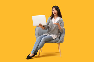 Beautiful Asian woman with laptop in grey armchair on yellow background