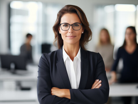 Mid-aged confident female entrepreneur posing smiling at camera whit their team in background in her white office.