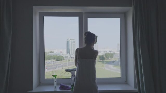 Silhouette Of Woman In Casual Home Clothes Washes Window Glass In Apartment On High Floor Of Residential Building Rear View